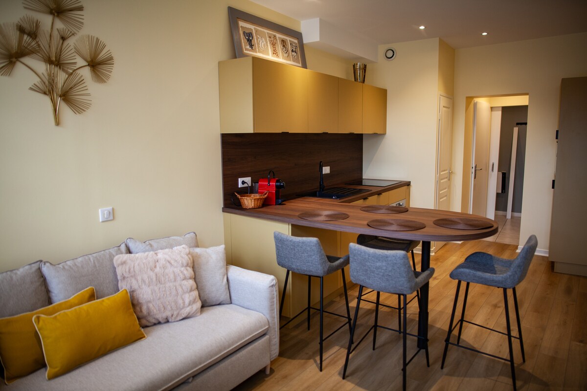A modern kitchen space features golden cabinetry and a dark wood countertop. A round wooden dining table with three high stools is positioned near a comfortable sofa adorned with cushions. Soft lighting enhances the warm tones of the walls and furniture.