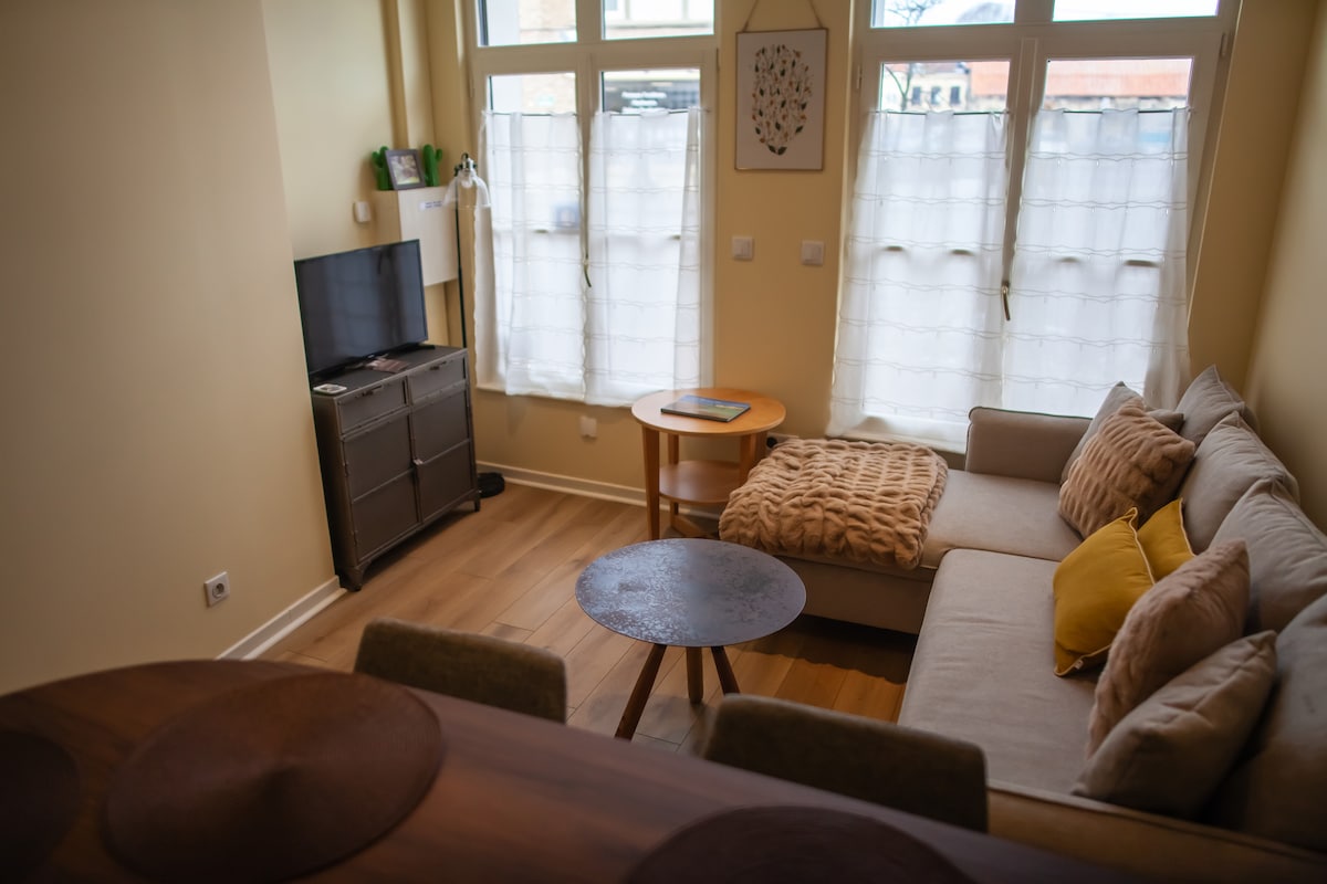 A cozy living space features a spacious sofa adorned with cushions, complemented by natural light from large windows. A small round table and a dark wooden TV stand enhance the room's functionality. Soft earth tones and a textured throw blanket contribute to a relaxed atmosphere.