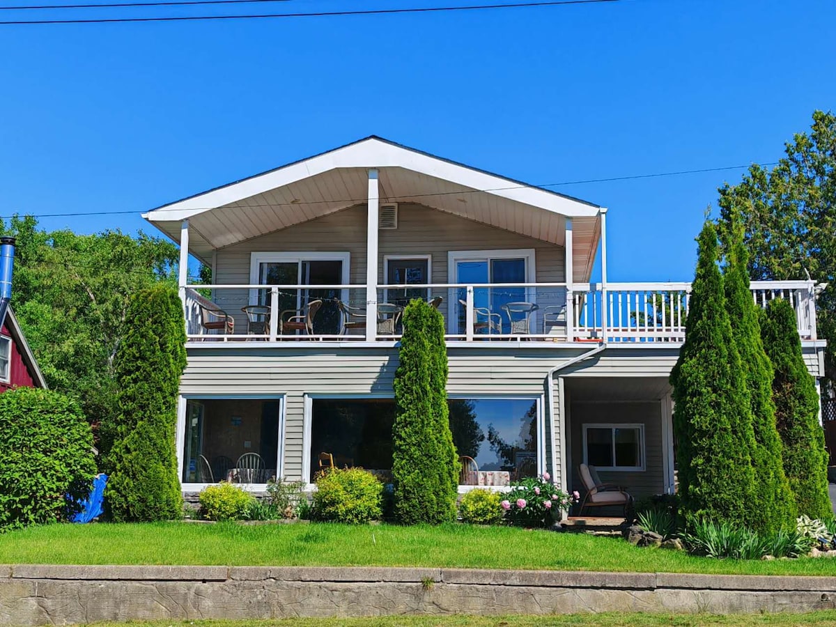A large two-story home is presented with a covered upper deck, featuring seating areas for relaxation. The front yard is landscaped with greenery and flowers, while large windows allow natural light to fill the interior. This inviting structure is set against a clear blue sky.