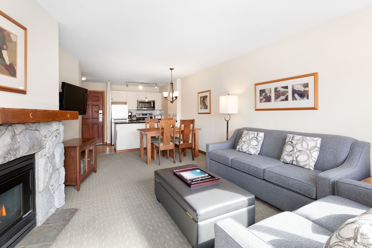 The living area features a gray sectional sofa adorned with decorative pillows facing a stone fireplace. A dining table with wooden chairs is positioned nearby, which connects to the fully equipped kitchen visible in the background. Natural light fills the space through a window, enhancing the welcoming atmosphere.