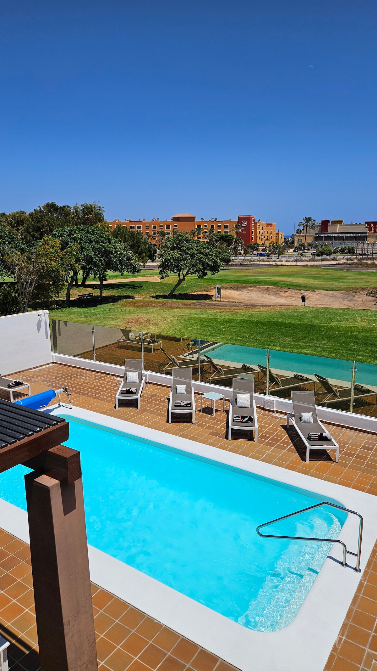 The image captures a private heated swimming pool with a tiled border and stairs leading in. Surrounding the pool are six padded deckchairs, and a glass fence provides an unobstructed view of the manicured golf course and vibrant buildings in the distance under a clear blue sky.