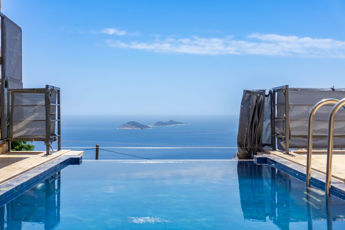 An infinity pool is visible, seamlessly merging with the horizon. The tranquil water reflects the clear blue sky and offers views of distant islands in the Mediterranean Sea. Lush landscaping frames the pool area, enhancing an inviting outdoor experience.