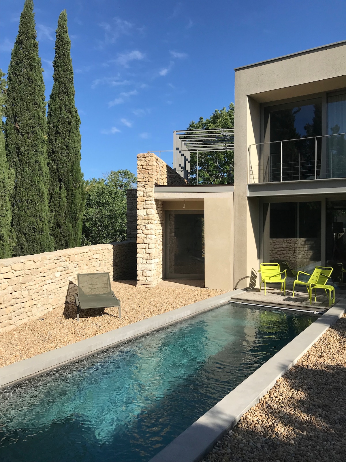 A contemporary swimming pool is surrounded by a pebbled courtyard, accompanied by tall cypress trees and a stone wall. Modern seating in vibrant yellow is arranged adjacent to the pool, inviting relaxation. Natural light brightens the exterior, showcasing clean lines of the structure.