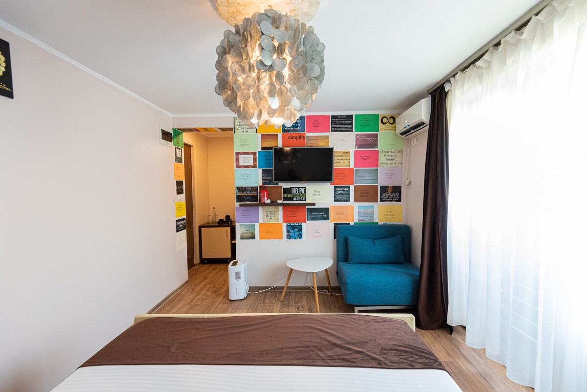 A well-lit room features a spacious bed with a simple white bedspread. A vibrant colored wall displays a variety of framed quotes. A cozy blue sofa and a round coffee table are positioned beside a window, allowing natural light to fill the space.