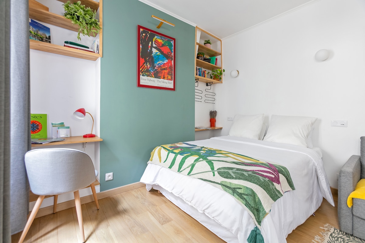 A cozy sleeping area features a spacious bed dressed in white linens, accented by a colorful throw blanket. Shelves above the bed display books and decorative items. A small desk with a lamp and a comfortable chair are positioned to the side, creating an inviting atmosphere.
