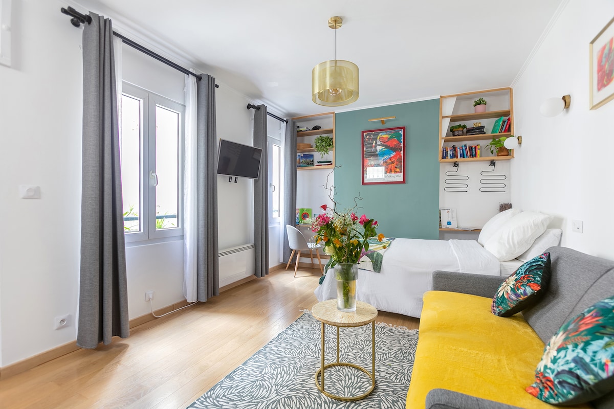 A cozy living area features a comfortable gray sofa and a vibrant yellow accent chair. Natural light filters through two large windows with gray curtains. A small table holds a floral arrangement, complementing the artwork on the walls and the bookshelf filled with books.