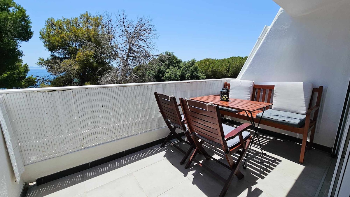An outdoor terrace features wooden furniture, including a rectangular table and four folding chairs. A cushioned bench is positioned against the wall. The backdrop includes lush greenery and a clear blue sky, providing an inviting space for relaxation.