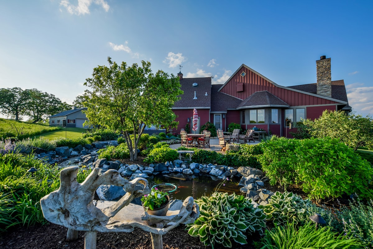 The exterior of the home is surrounded by lush greenery and vibrant landscaping, featuring a stone pond framed by diverse plants. A stone sculpture with a plant pot is visible in the foreground, while comfortable seating and a cozy atmosphere can be observed on the patio area.