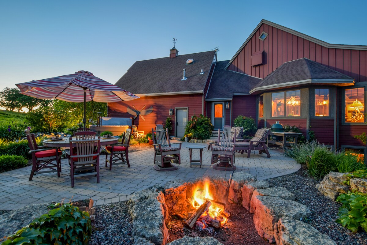 An inviting outdoor space features a stone fire pit with a warm fire, surrounded by several wooden lounge chairs. A patio table with umbrella provides a shaded dining area, while lush greenery and blooming flowers enhance the tranquil setting against the backdrop of a rustic home.