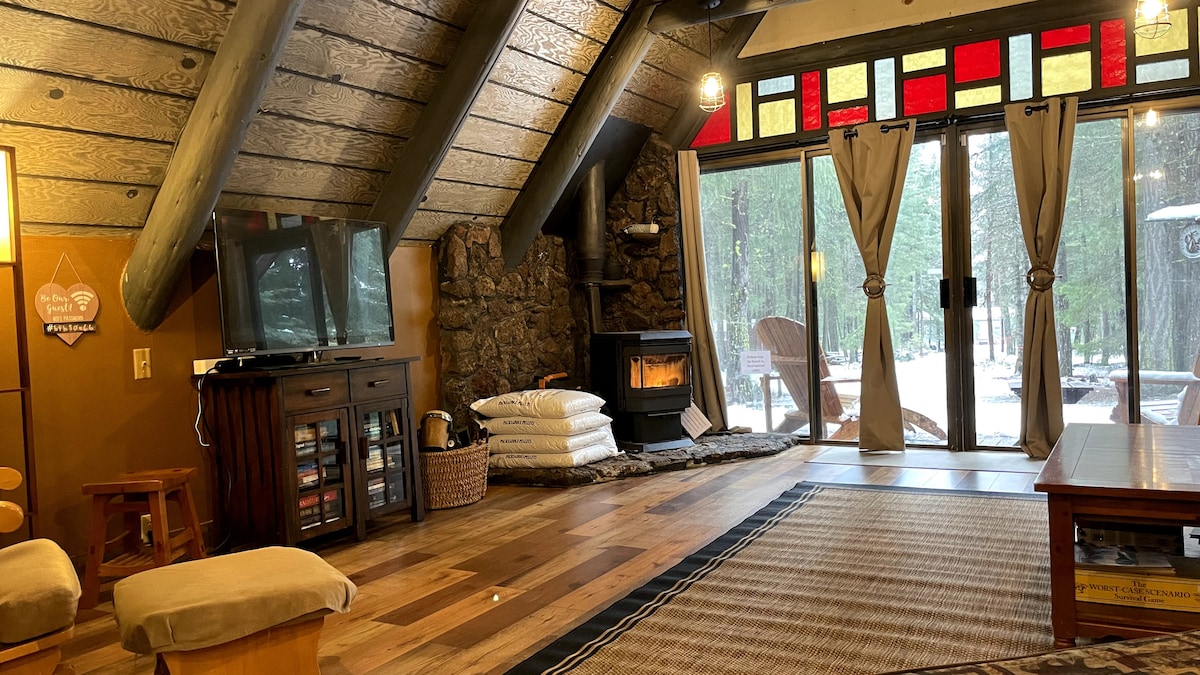 An inviting living area features a stone fireplace surrounded by comfortable seating. Large glass doors open to a forest view, with red and yellow stained glass accents above. A wooden coffee table and a woven basket are positioned on a wooden floor, enhancing the cabin's rustic charm.