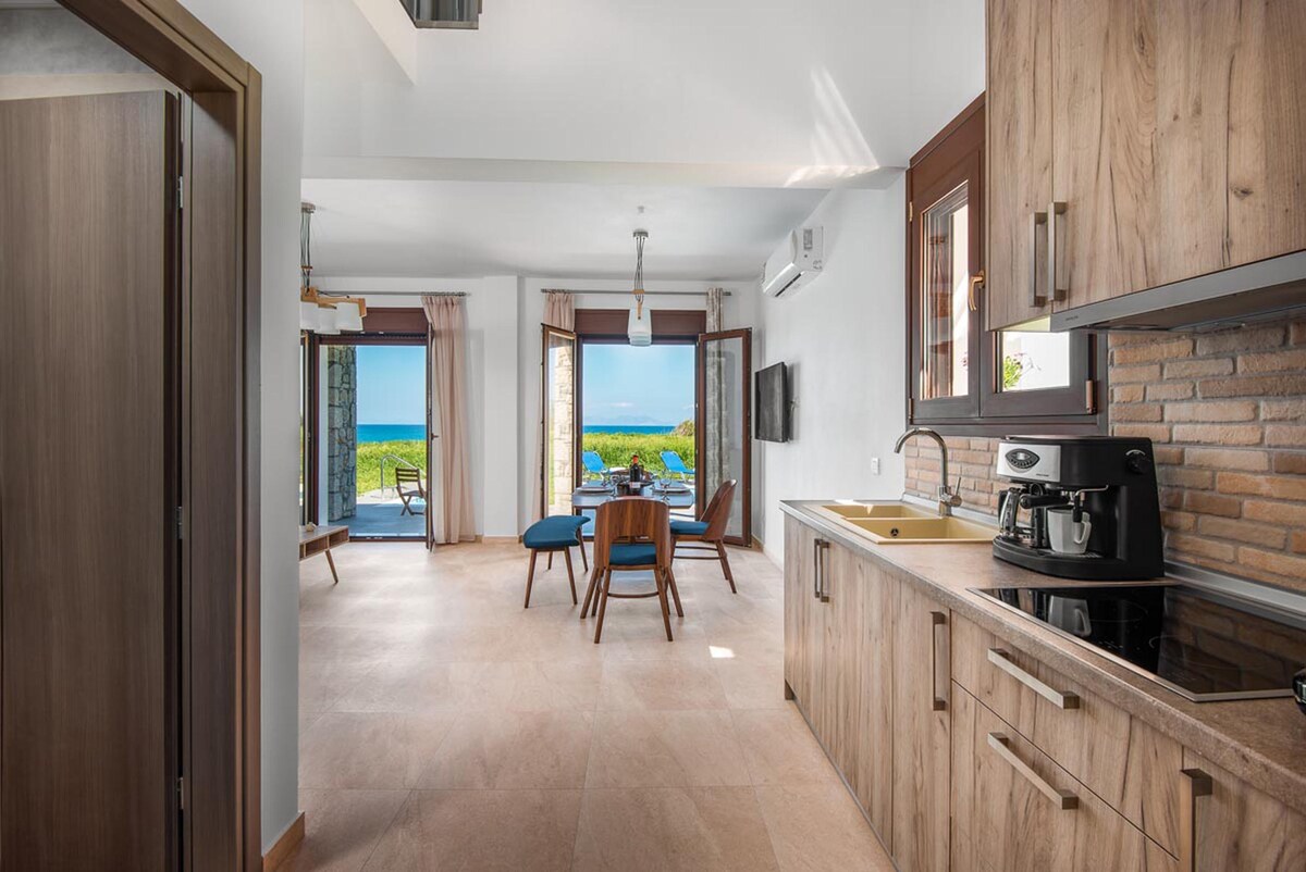 An open plan living area features a modern kitchen with sleek cabinetry, a dining table, and cozy seating. Large glass doors offer a view of the sea and garden, allowing natural light to illuminate the space. Air conditioning units are visible, ensuring comfort indoors.