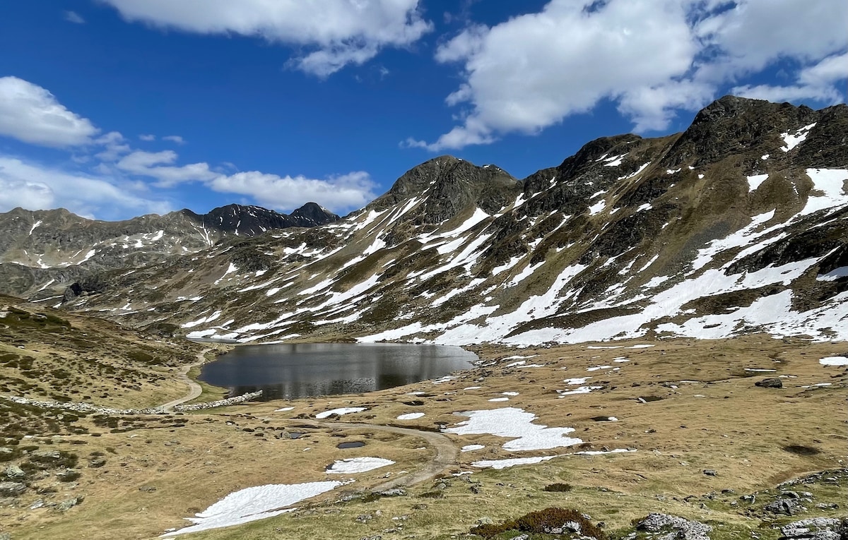 A serene mountain landscape features a calm lake set against a backdrop of snow-capped peaks. The foreground showcases rolling meadows with patches of snow, under a clear blue sky dotted with clouds.