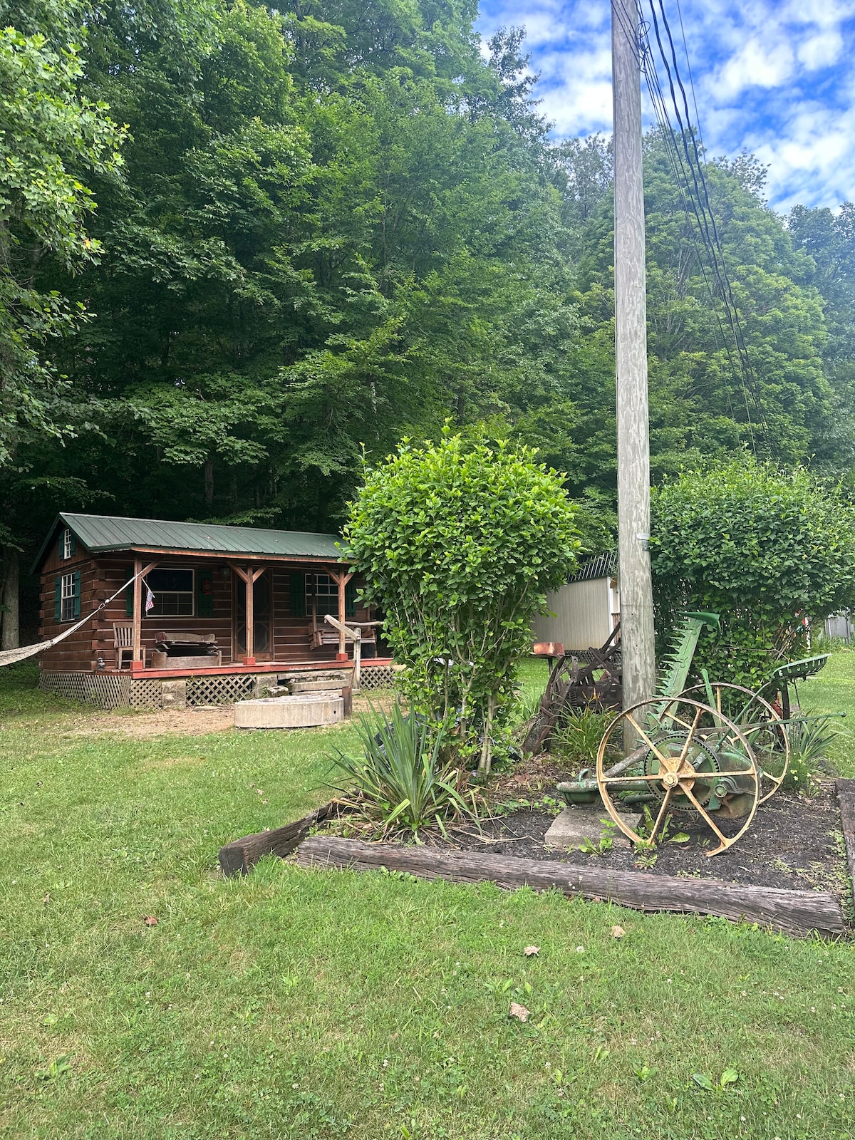 A rustic cabin is nestled among tall trees, showcasing a covered porch and a green roof. A stone fire pit is visible in front, surrounded by well-maintained greenery and decorative elements, including an antique wheelbarrow, enhancing the natural surroundings.