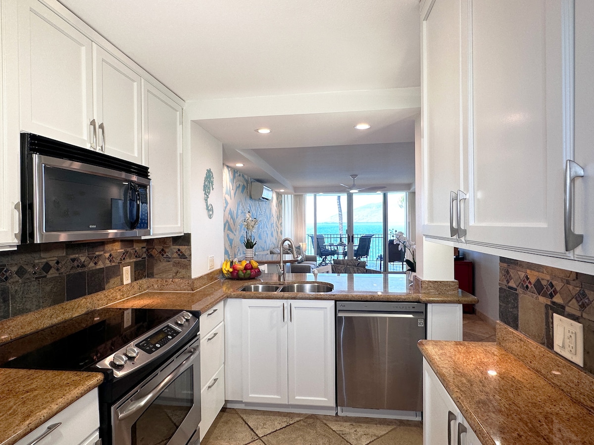 The modern kitchen features stainless steel appliances, including a gas stove and microwave, set against light cabinetry and granite countertops. Tile flooring extends throughout the space. A view of the ocean can be seen through the adjacent living area, providing natural light and a welcoming ambiance.