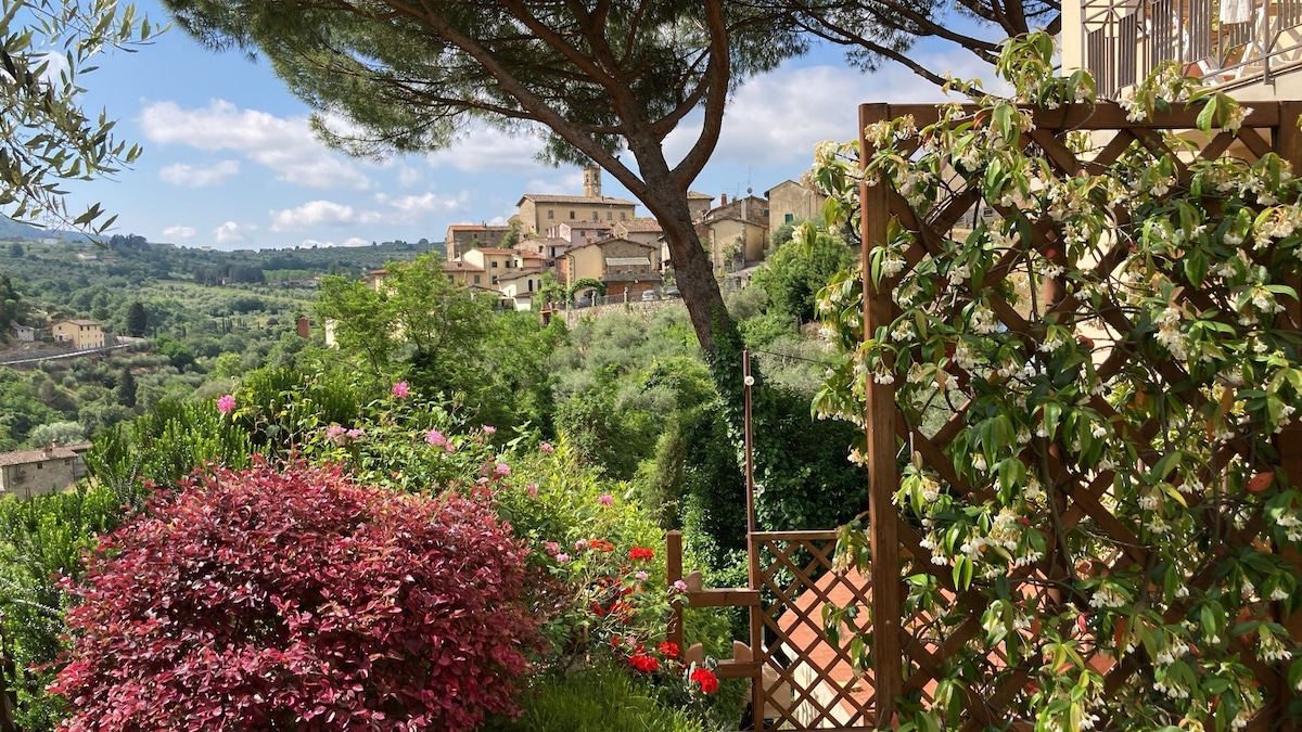 A lush garden area is visible, featuring vibrant flowers in various colors. A wooden trellis can be seen, partially covered with green foliage. In the background, hills dotted with quaint houses and a clear blue sky enhance the serene atmosphere.