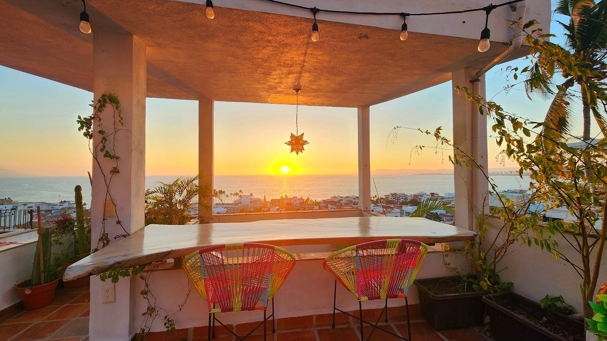 An outdoor terrace offers an unobstructed view of the sunset over the ocean. A wooden table is flanked by two colorful chairs, surrounded by plants. Soft lighting is provided by hanging bulbs, enhancing the tranquil ambiance as the sun sets on the horizon.