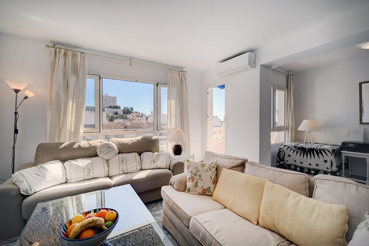 The living room features a spacious layout with two large sofas, one adorned with decorative pillows. A coffee table holds a bowl of fresh fruit. Large windows let in natural light and offer views of the surrounding area.