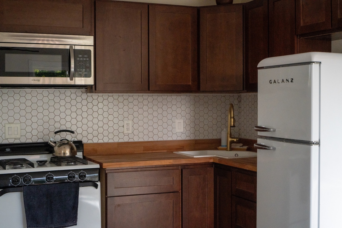 A modern kitchen features wooden cabinetry and a sleek countertop. Stainless steel appliances, including a microwave and gas stove, are complemented by a retro-style refrigerator. A stylish hexagon backsplash adds texture, while a kettle is placed on the stove, ready for use.