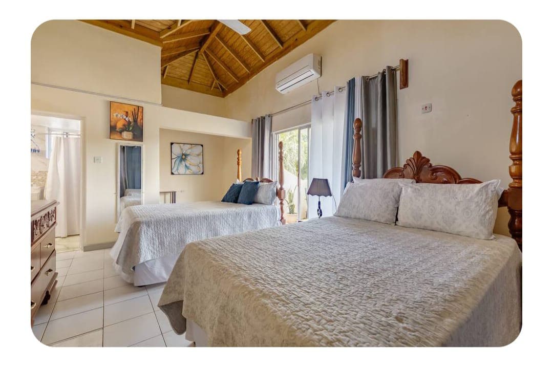 The upper room features a high ceiling with wooden beams, two spacious double beds adorned with light bedding. Large windows allow natural light to fill the space, and a private balcony is visible, offering views of the surrounding area.