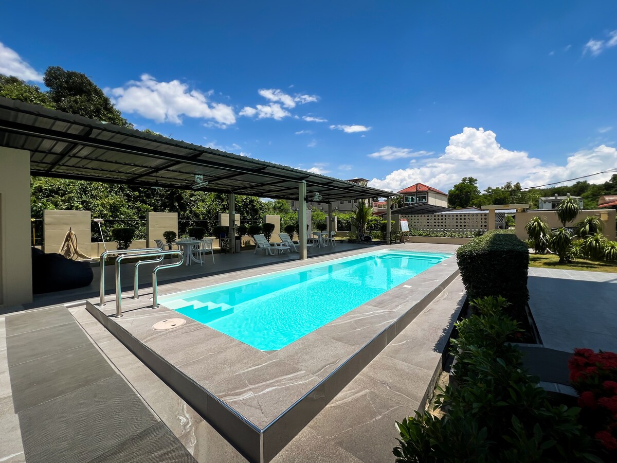 A sparkling pool is framed by a sleek stone deck, with lounge chairs arranged nearby. Lush greenery surrounds the area, complemented by a shaded seating space beneath a roof. Clear blue skies and fluffy clouds create an inviting backdrop.