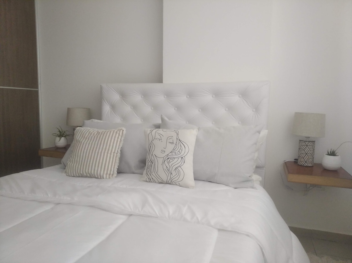 The sleeping area features a spacious bed with a white tufted headboard, complemented by light grey and patterned decorative pillows. Nightstands with small lamps are positioned on either side, and a small potted plant adds a touch of greenery to the room.