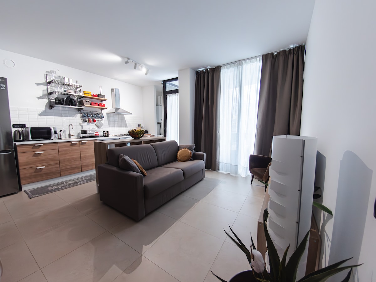 A modern living area is showcased with a spacious, neutral-toned sofa facing an open kitchen. Natural light filters through sheer curtains, illuminating a sleek kitchen design featuring shelves with kitchenware and appliances. A potted plant adds a touch of greenery to the space.