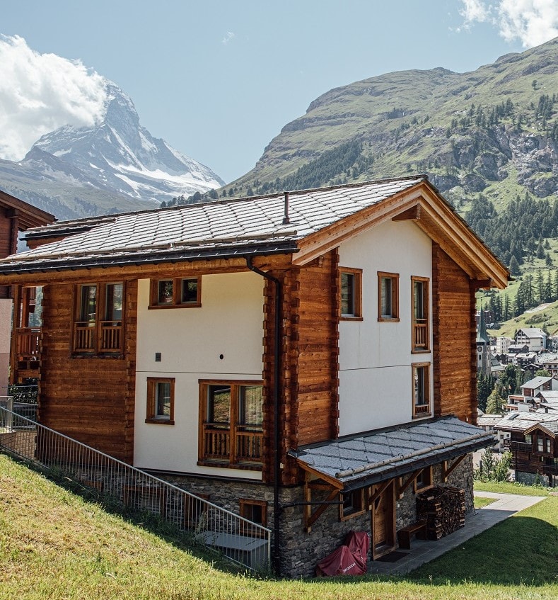 A modern wooden chalet stands within a scenic mountainous backdrop. The structure features a stone base and multiple large windows that offer views of the surrounding landscape. A sloped green area leads up to the entrance, enhancing the natural setting.