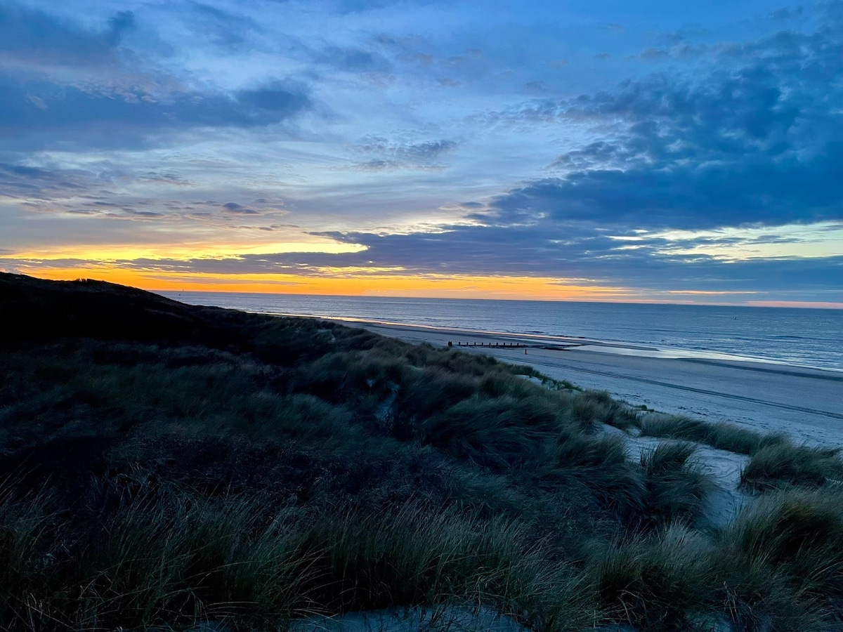 A serene beach scene is captured during twilight, featuring soft sand dunes outlined by gentle grasses. The horizon showcases a gradient of colors as the sun sets, illuminating the sky with shades of orange and purple over calm waters. A distant wooden walkway is visible along the beach.