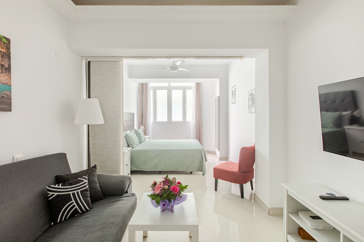 A modern living area features a dark gray sofa with patterned cushions and a white coffee table displaying a vase of flowers. Adjacent, a spacious bedroom is visible, with a large bed covered in green and white bedding. A window allows natural light to enter.
