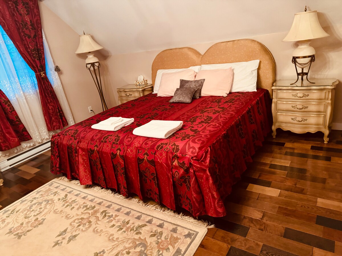 A cozy sleeping area features a spacious bed with a rich red bedspread adorned with intricate patterns. Two bedside tables with lamps are positioned beside the bed. A large area rug lies beneath, and a window with light, flowing drapes allows natural light to enter.