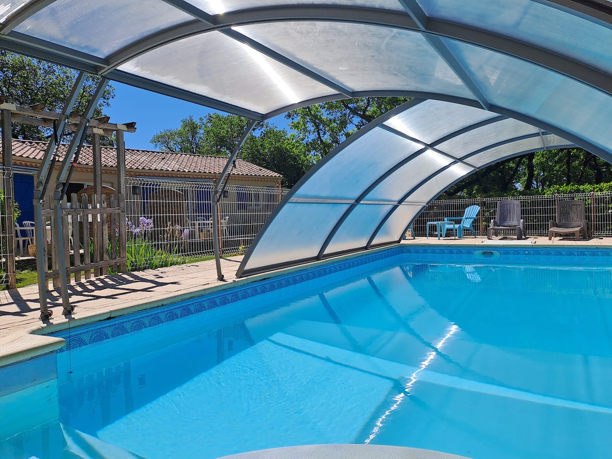 A spacious outdoor pool is enclosed under a clear canopy, revealing bright blue water that glistens in the sunlight. Surrounding the pool are several lounge chairs, a wooden pergola, and lush greenery, creating a serene setting for relaxation.