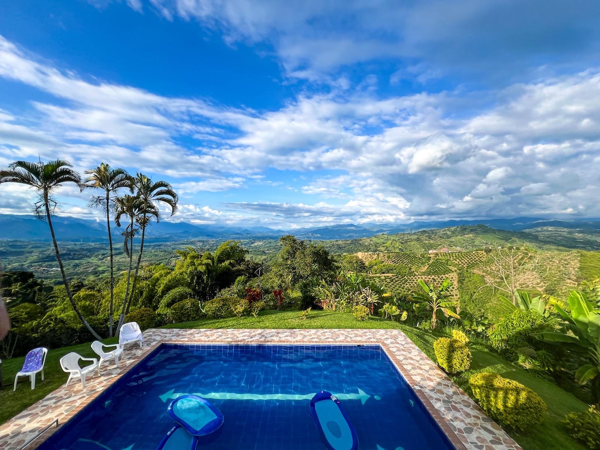 A sparkling pool is framed by lush greenery, showcasing a panoramic view of rolling hills and cloudy skies. Lounge chairs are positioned nearby, inviting relaxation while taking in the tranquil landscape of the coffee region.
