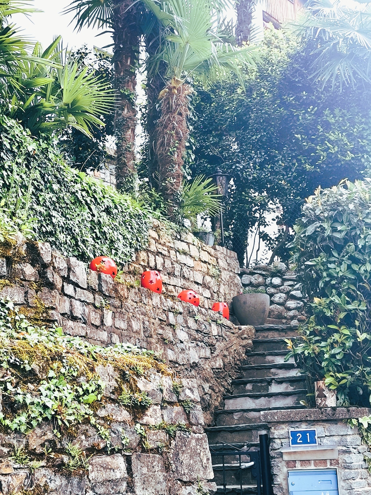 A stone pathway lined with lush greenery leads up toward a stairway. Bright red pots are placed along the steps, adding a touch of color against the natural surroundings. A house number plaque is visible near the base of the stairs.
