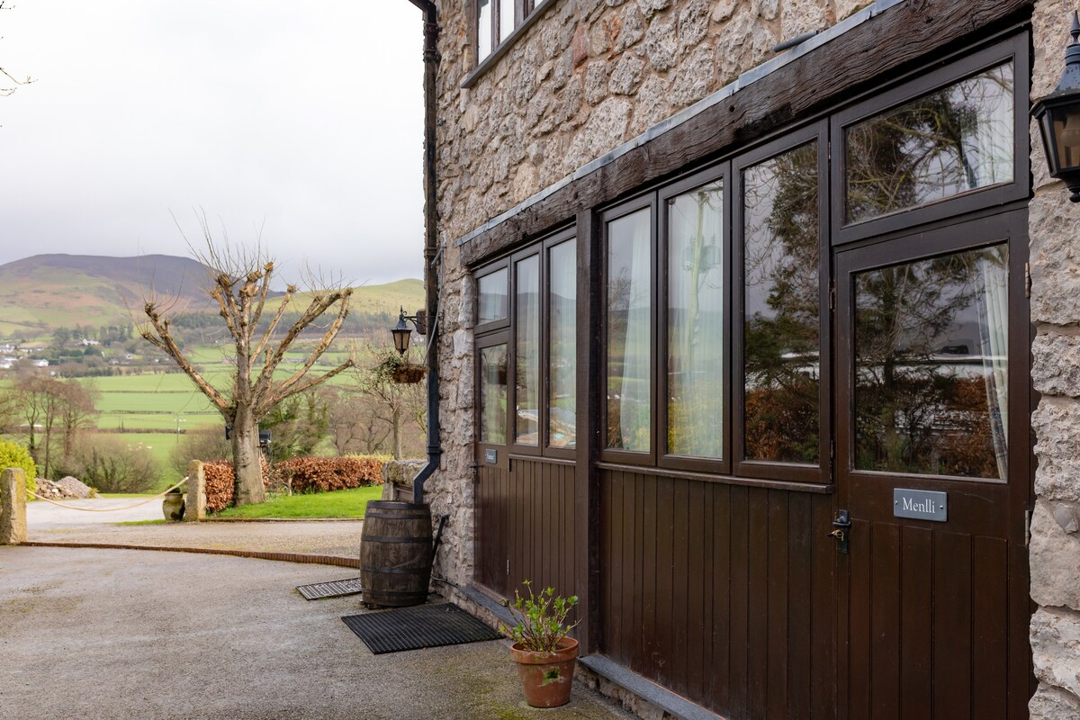 A stone exterior is featured, showcasing large windows that provide views of the surrounding landscape. A small potted plant adds a touch of greenery, while a barrel is placed nearby. The pathway leading to the property is visible, along with rolling hills in the distance.