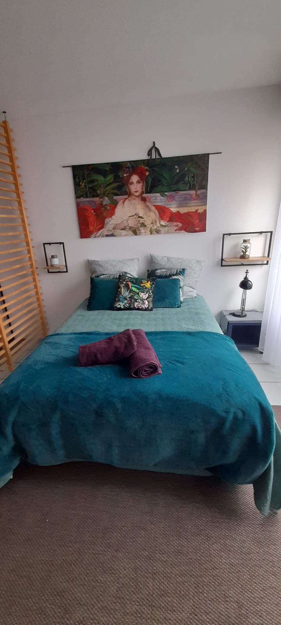 A well-decorated sleeping area features a spacious bed dressed in teal bedding. Art is displayed above the bed, and decorative pillows are arranged on top. A nightstand holds a lamp, with towels thoughtfully placed on the bed, adding to the room's functionality.