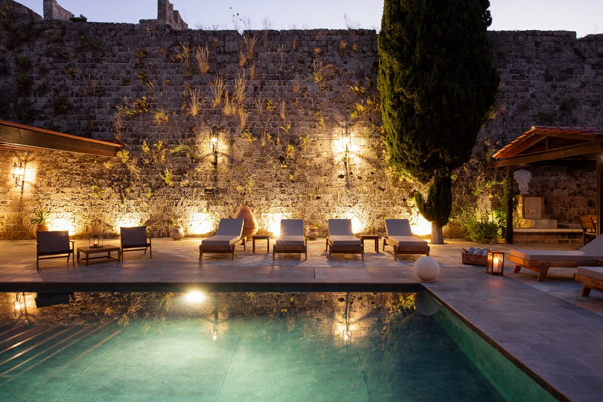 The serene outdoor space features a swimming pool surrounded by comfortable sunbeds and gently glowing lights. A stone wall provides privacy, with lush greenery adding a touch of nature. The inviting atmosphere is enhanced by warm lighting, creating a relaxing environment for guests.