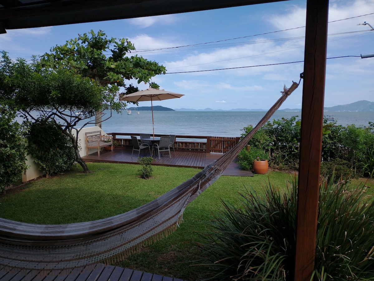 A hammock is suspended under a wooden structure, offering a view of the tranquil waters. A large shaded patio with seating and a dining table is visible, surrounded by lush greenery and a well-manicured lawn.