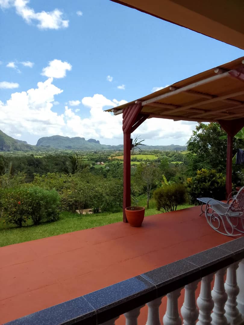 A spacious terrace is seen overlooking lush greenery and panoramic mountain views. The area is shaded by a roof, and outdoor seating is arranged to enjoy the scenery. Bright blue skies with scattered clouds enhance the tranquil ambiance of the space.