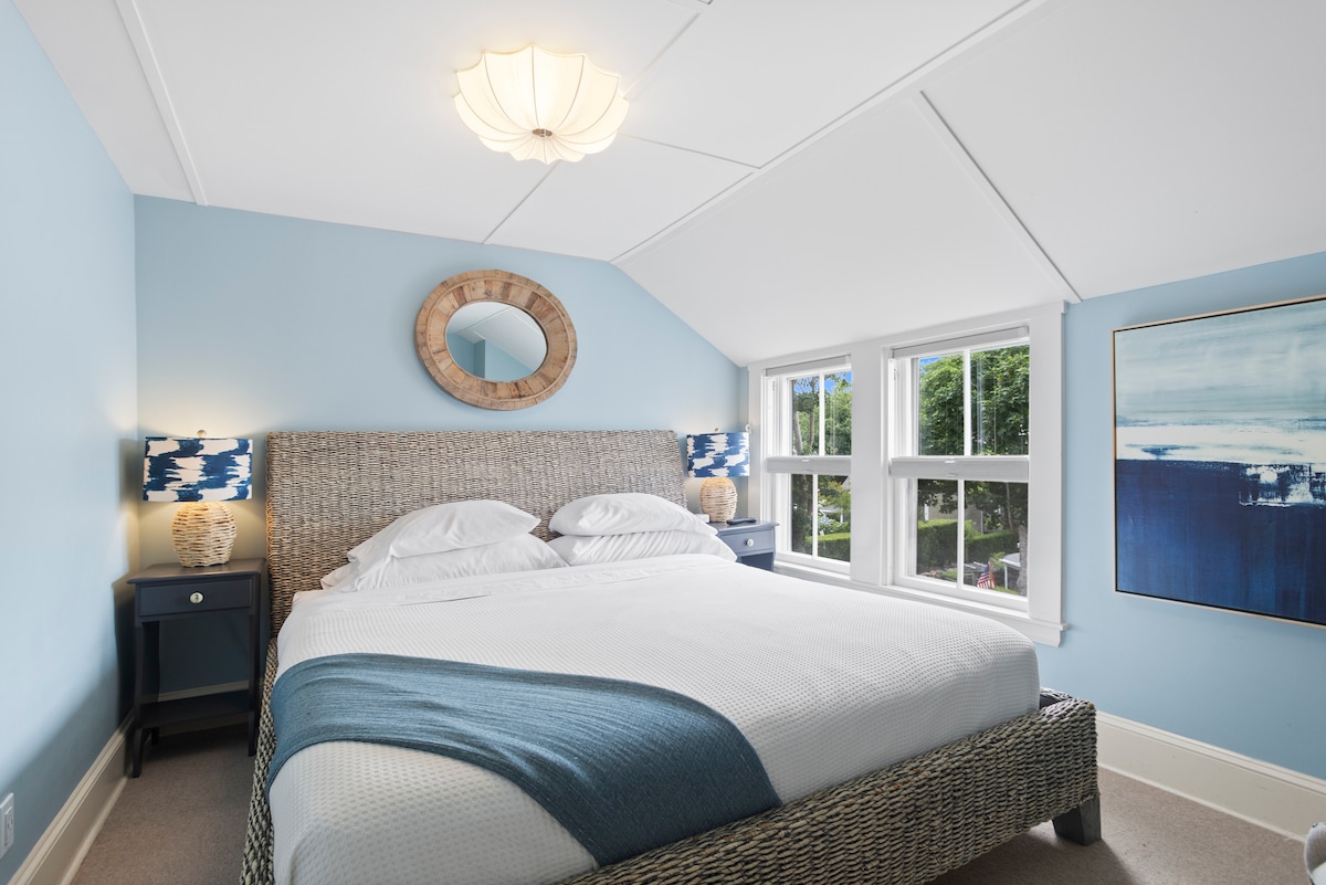 A well-lit bedroom features a spacious bed with light linens and a soft blue throw. Natural light enters through two large windows, framed by white trim. A round mirror and bedside tables with decorative lamps add a touch of charm to the serene atmosphere.