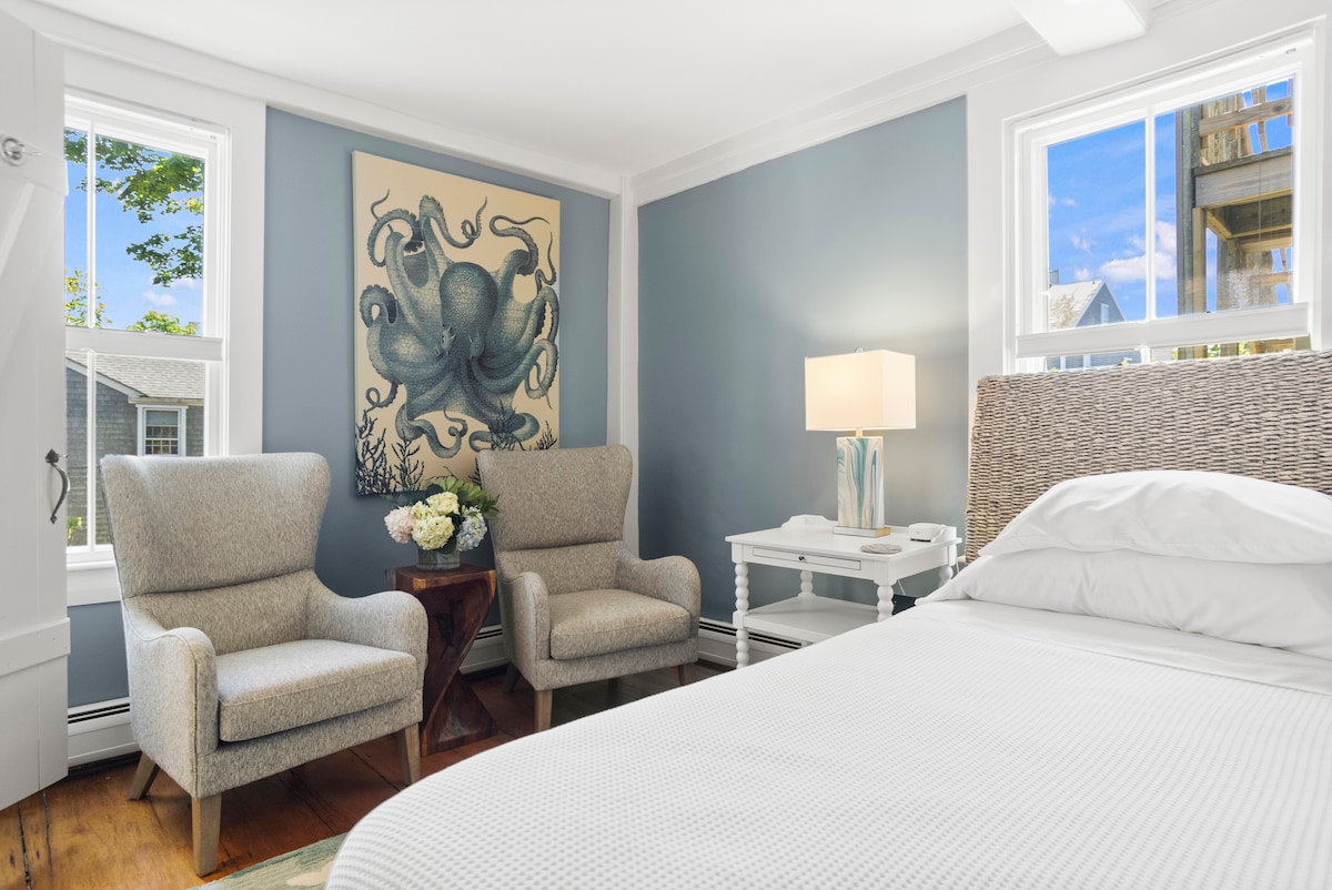A well-lit room features a spacious bed with a light-colored coverlet. Two comfortable armchairs are positioned near a side table. A large piece of wall art depicting an octopus adds character, while windows provide natural light and a view of the garden.