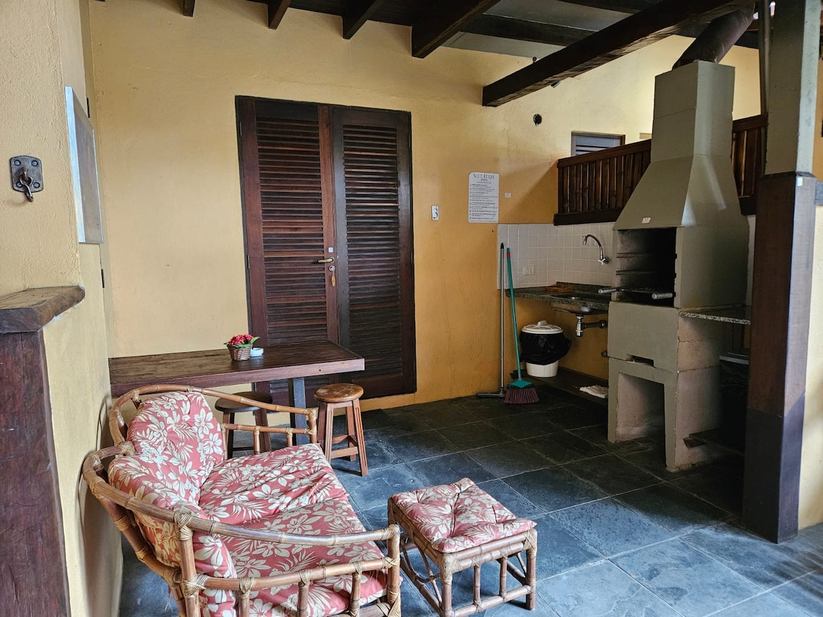 An indoor seating area features a wicker chair and ottoman, coupled with a small table. A rustic kitchen area is equipped with a grill and basic countertop appliances. Natural light illuminates the space, highlighting the textured stone flooring and warm yellow walls.