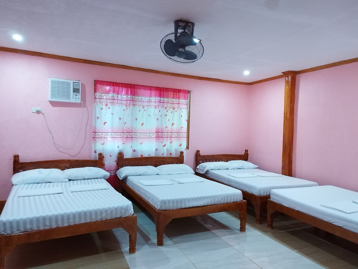 A cozy sleeping area is presented with four spacious beds arranged in a clean layout. Soft, white bedding contrasts with the light pink walls. A ceiling fan is visible, and natural light filters through a window covered by a floral curtain.