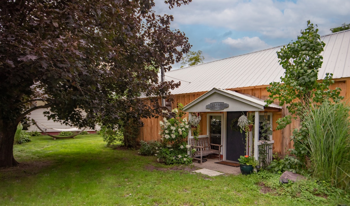 The exterior of a rustic cottage is displayed, featuring a covered entrance with a welcoming sign. Flanking the entrance are flower arrangements and greenery. A small bench sits beside the door, and a grassy lawn extends around the cottage, complemented by shade from nearby trees.
