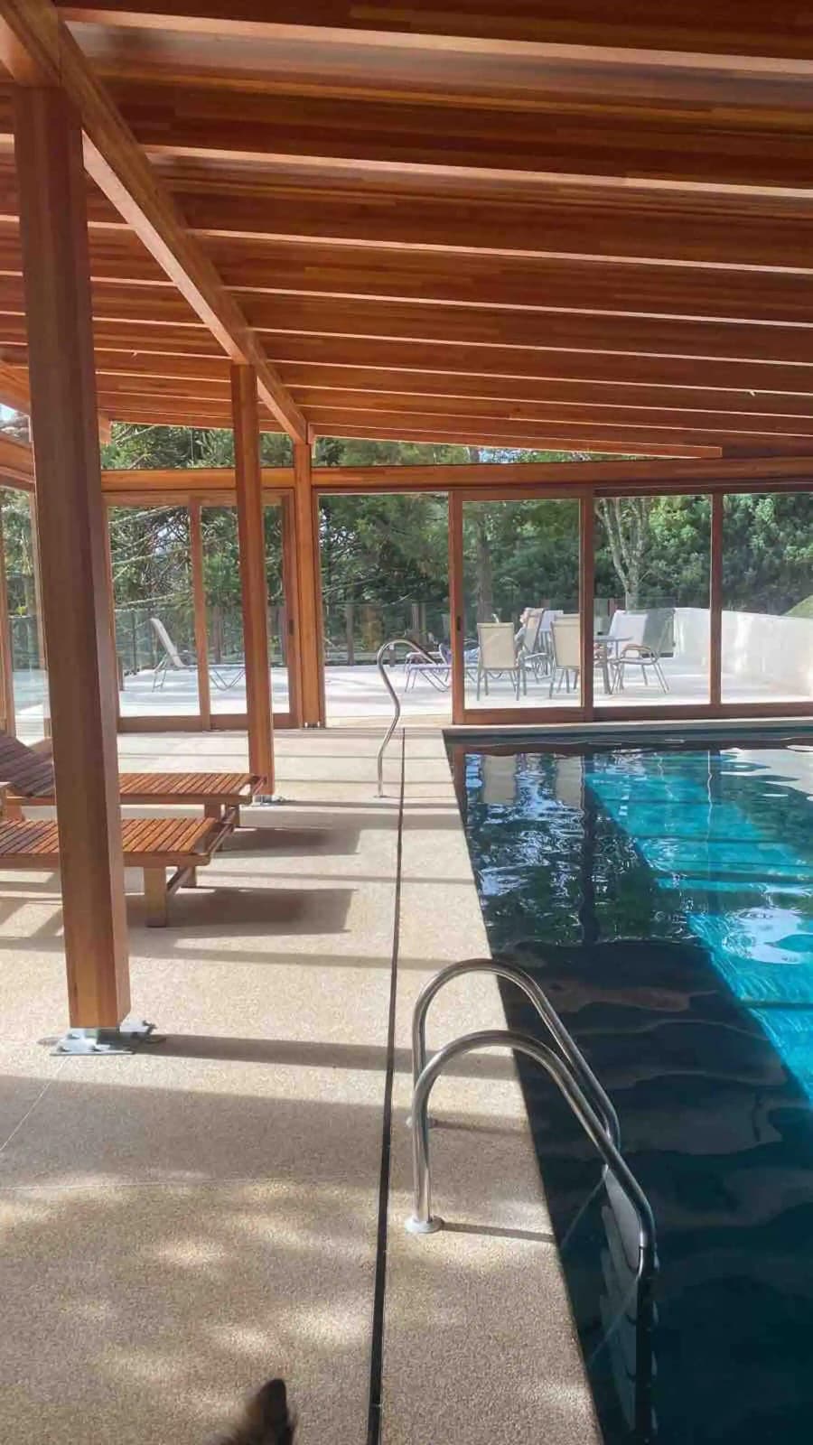 A covered outdoor area features a swimming pool with clear water, surrounded by large glass panels. Several lounge chairs are positioned nearby on a smooth surface, offering a calm space to relax. Comfortable seating can be seen in the background, emphasizing a laid-back environment.