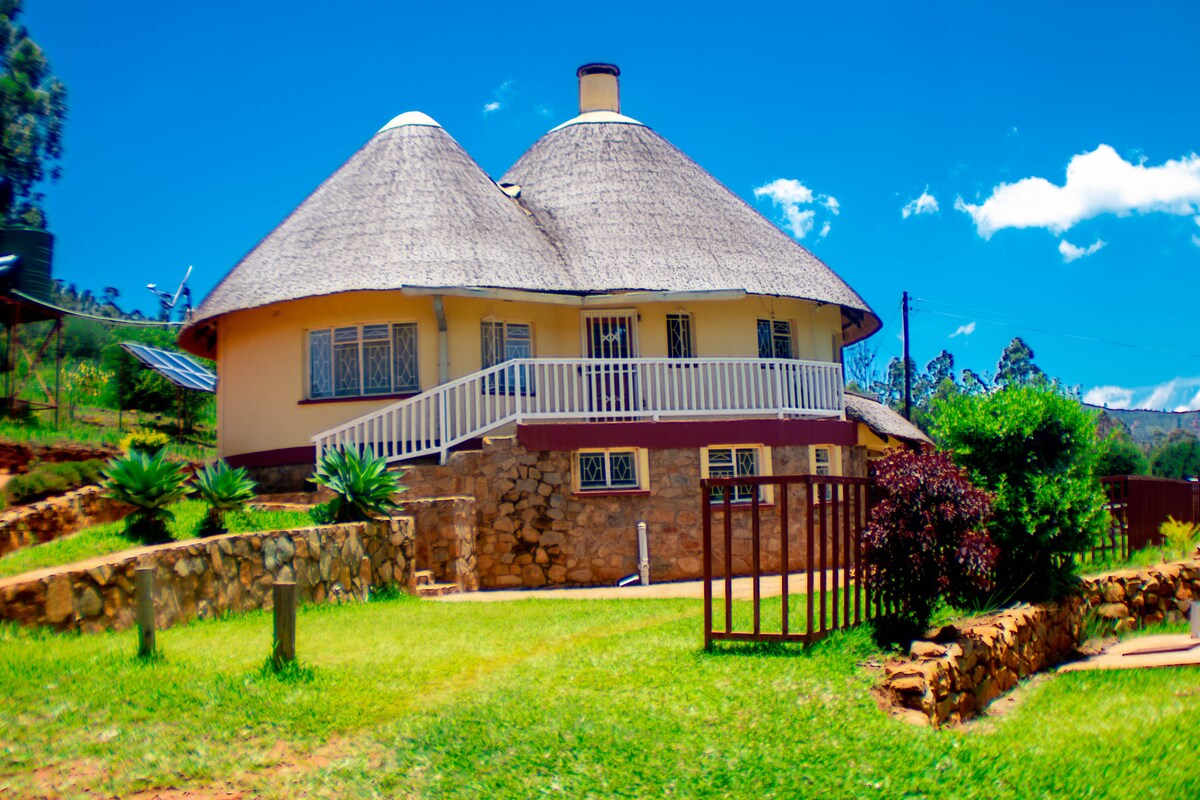 A round-shaped cottage with a thatched roof is surrounded by well-maintained gardens. The exterior features stone walls and a white railing on the upper balcony. Lush green grass and decorative plants contribute to the inviting landscape under a clear blue sky.
