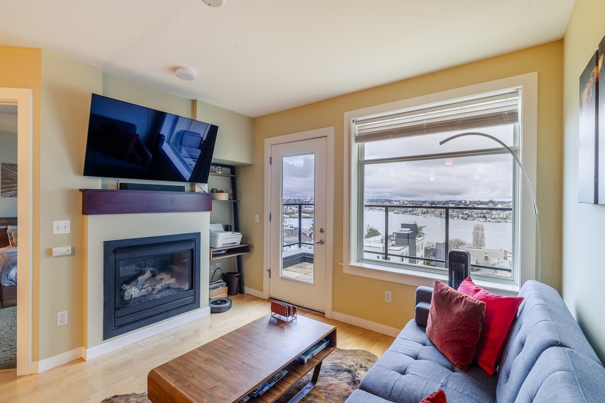 The living room features a cozy sofa adorned with red accent pillows. A mounted flat-screen TV overlooks a coffee table, while large windows provide a view of Lake Union and the surrounding landscape. A door leads to an outdoor patio space.