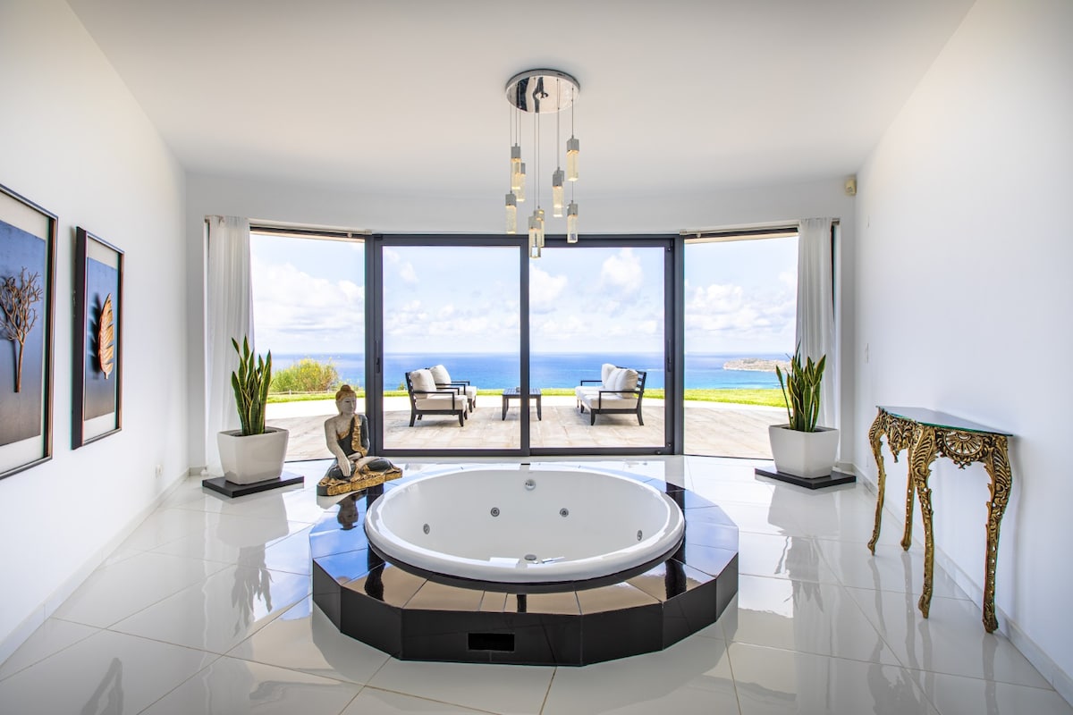 A sunlit room features a circular indoor Jacuzzi, surrounded by elegant white tiles. Large glass doors offer a clear view of the sea, while potted plants add a touch of greenery. A decorative table and artwork enhance the serene atmosphere.