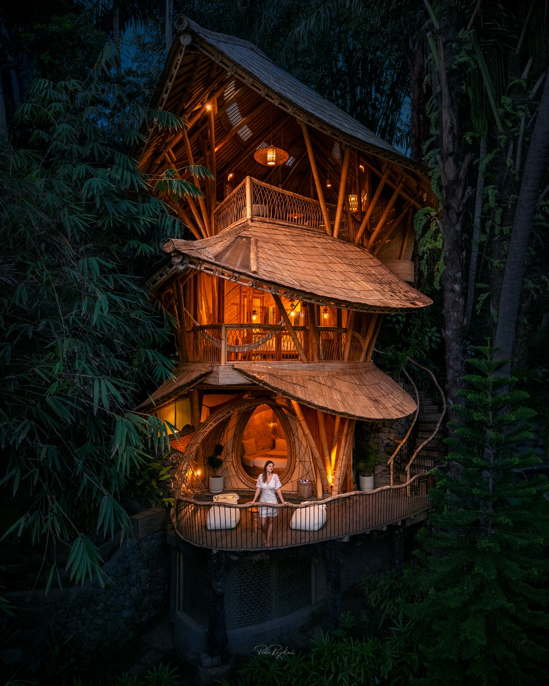 The bamboo house is illuminated warmly at dusk, showcasing its unique architecture with multiple levels. A spacious deck features lounge chairs surrounded by lush greenery, while the distinctive egg-shaped door of the second bedroom adds a focal point to the design.