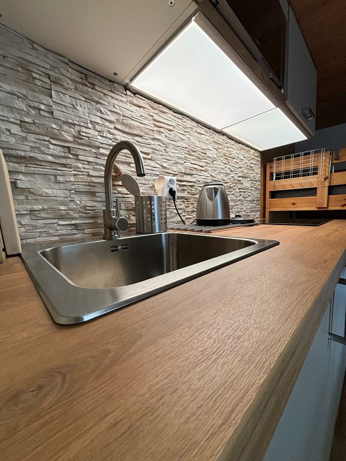 The kitchen features a polished wooden countertop with a stainless steel sink and modern faucet. The backdrop is adorned with textured stone tiling, and under-cabinet lighting enhances the workspace. A kettle and utensil holder are visibly positioned on the counter, contributing to the functionality of the area.