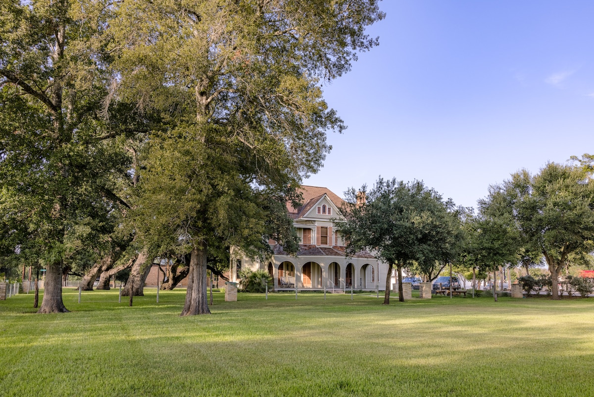 A historic late-Victorian home is nestled among mature oak trees, showcasing its charming architecture. The well-maintained grounds feature expansive green lawns, offering ample space for outdoor activities. The house boasts multiple levels and classic details, inviting exploration and enjoyment of the surroundings.